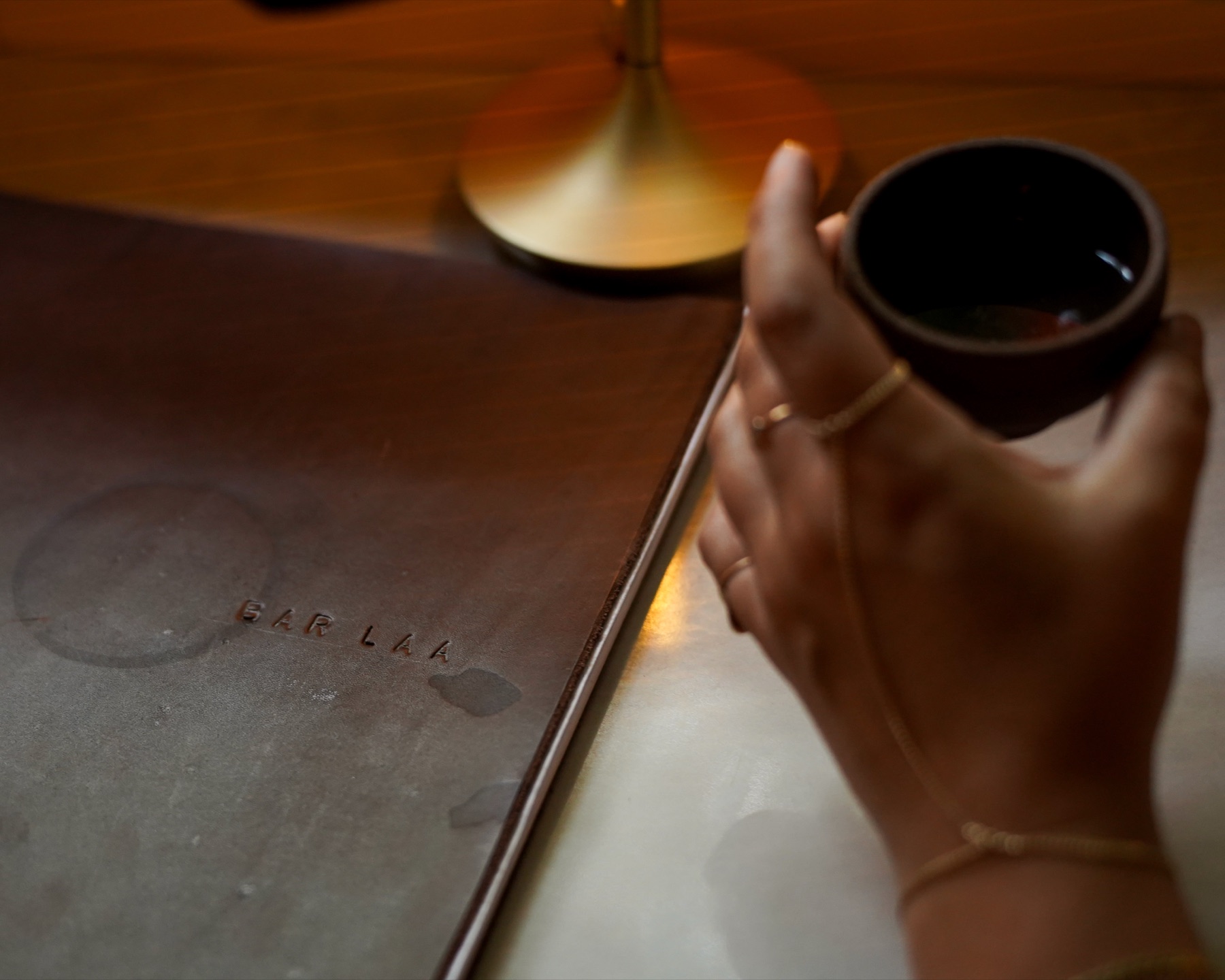 A hand cradles a small clay cup beside a leather Bar Laa menu, candlelight warm in the background